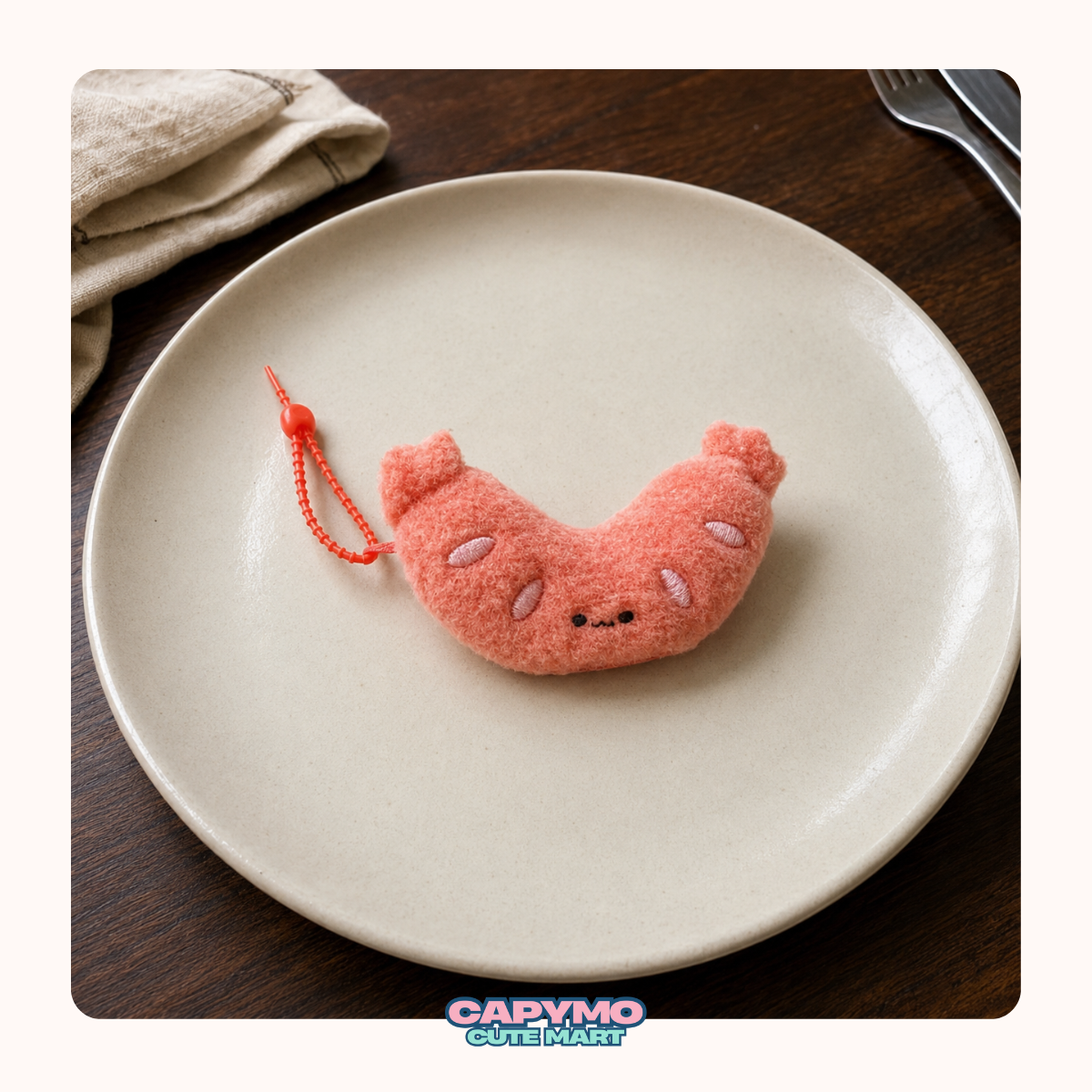 A small plush sausage-shaped keychain or charm in soft peach-pink with embroidered facial features and a red hanging string, placed on a round beige plate over a dark wood surface. A folded beige cloth and part of a fork are visible in the background.