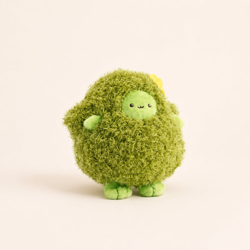 A cute, round green plush toy with a shaggy, moss-like texture, a small smiling face, and tiny green feet, standing against a solid off-white background.