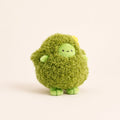 A cute, round green plush toy with a shaggy, moss-like texture, a small smiling face, and tiny green feet, standing against a solid off-white background.