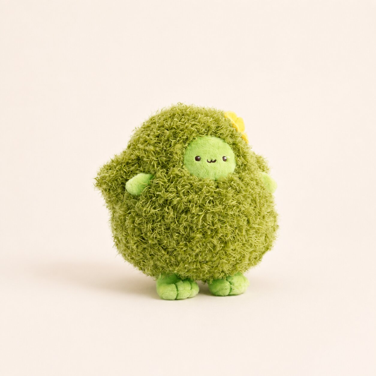 A cute, round green plush toy with a shaggy, moss-like texture, a small smiling face, and tiny green feet, standing against a solid off-white background.
