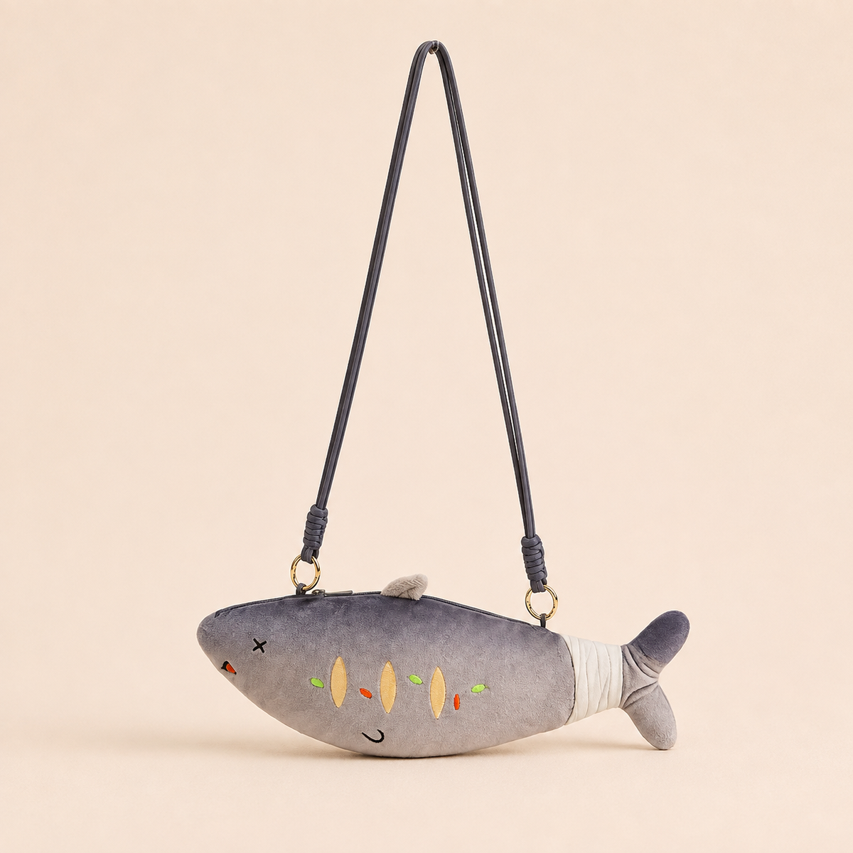 A plush fish-shaped shoulder bag in gray with embroidered details, a black strap, and gold-tone hardware, photographed against a solid pale beige background.