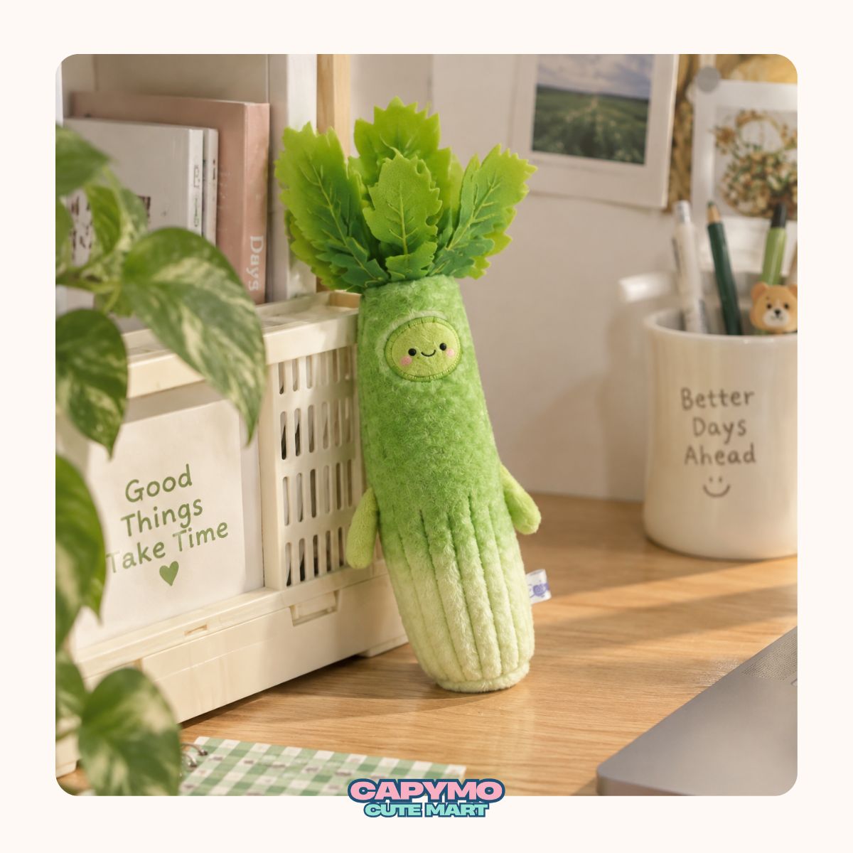 A cute plush toy shaped like a green pickle with leafy greens on top, sitting on a wooden desk in a styled room setting. The brand name 'Cadymo Cute Plush' is visible at the bottom of the image.