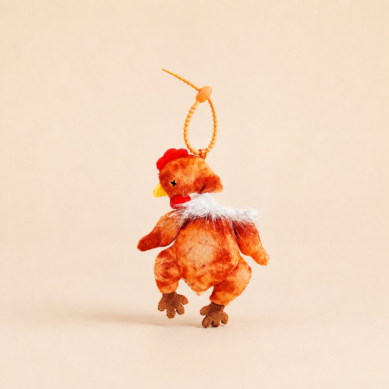 A small plush chicken keychain or bag charm with a red comb, yellow beak, brown body, and white fluffy neck detail, hanging from an orange cord on a plain beige background.