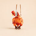 A plush chicken hanging ornament or decoration shown from the back, featuring a textured orange-brown body, red comb, white feather detail, and two hanging strings against a plain beige background.
