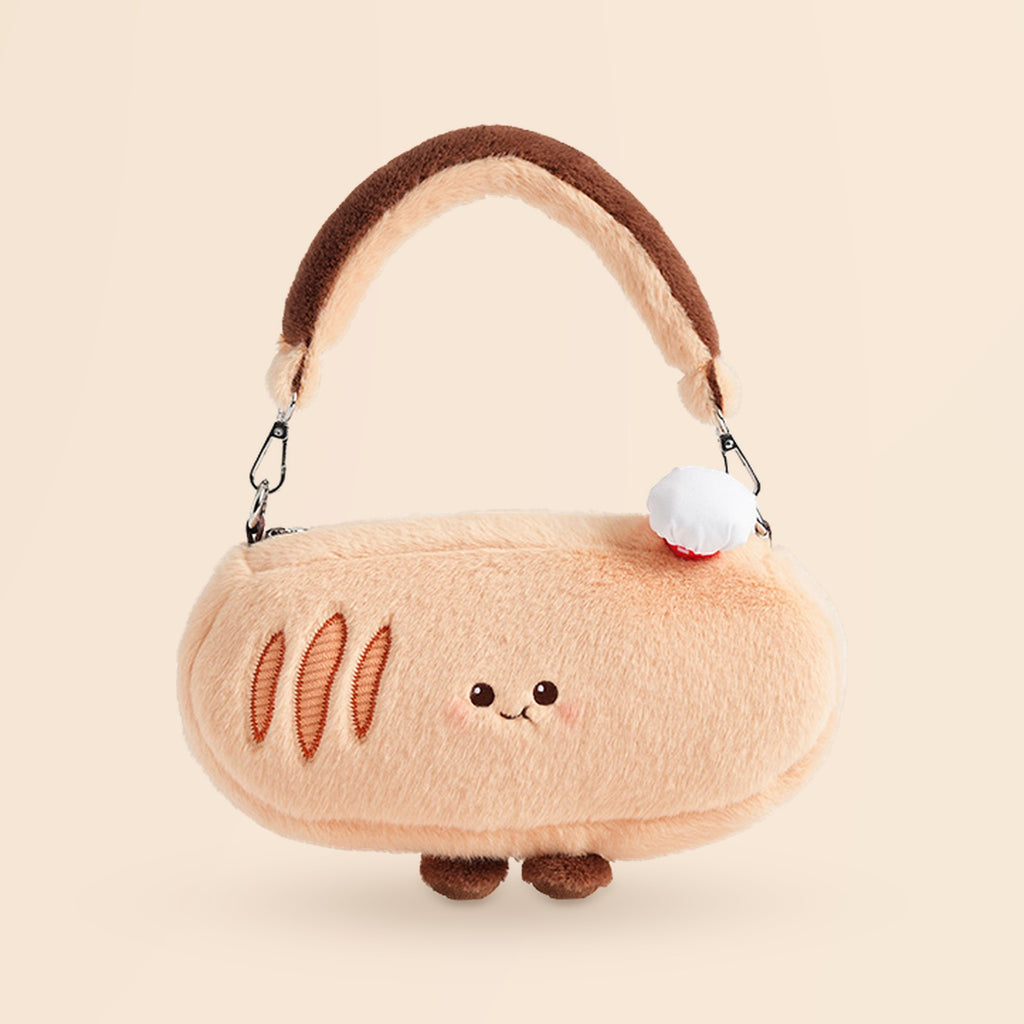 A plush bread-shaped handbag in a soft peach-beige color with embroidered crust details, a brown handle, small brown feet, and a white pom-pom accent, shown on a plain warm beige background.