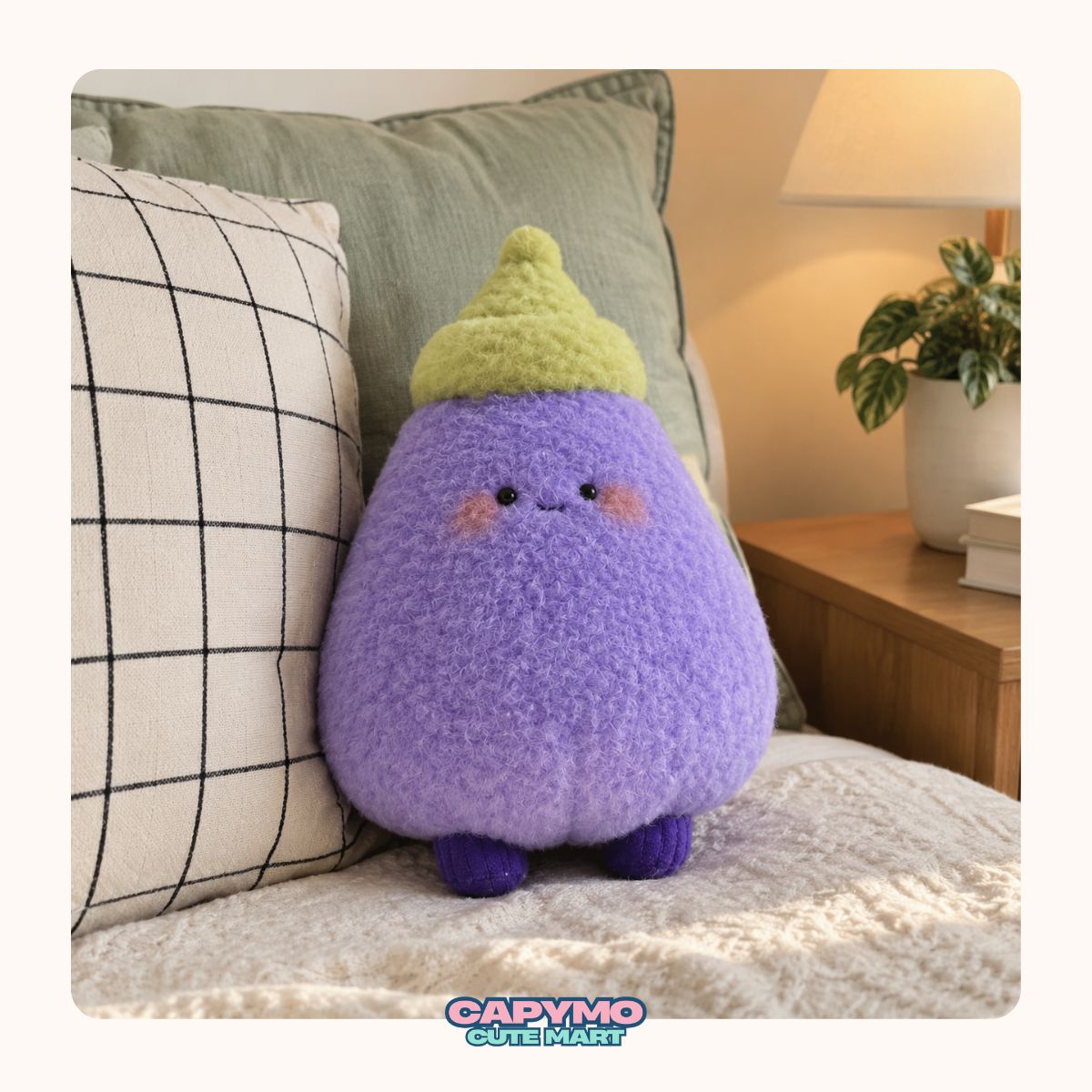 A cute plush eggplant toy with a fuzzy purple body, small dark eyes, pink cheeks, and a yellow-green pointed hat, sitting on a bed with pillows in a warm bedroom setting.