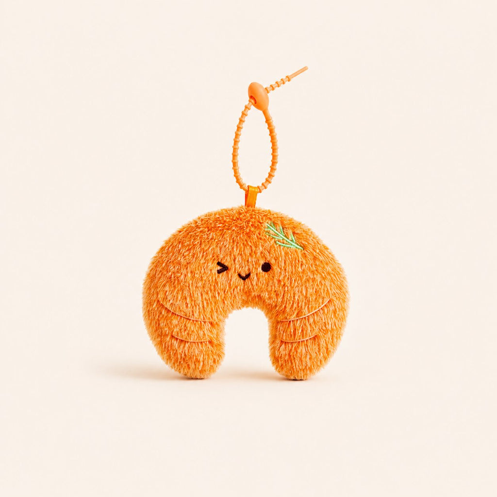 An orange plush crescent-shaped keychain or bag charm with a small green leaf detail, embroidered face, and a beaded orange loop at the top, shown on a plain cream background.