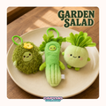 Three cute plush vegetable characters labeled 'GARDEN SALAD' are displayed on a beige plate over a wooden surface. The set includes a fuzzy green mossy character with a yellow flower, a green cucumber-shaped plush with leafy top, and a round light green radish character with pink cheeks.