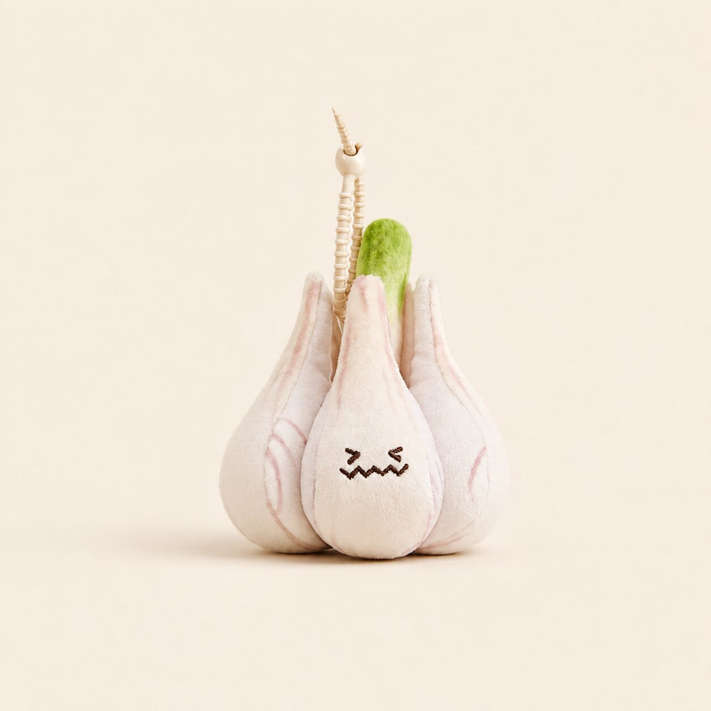 A plush garlic toy with a stitched frowning face, featuring pale pink and off-white fabric cloves, a green stem, and a small wooden bead keychain attached at the top, set against a solid cream background.