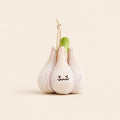 A plush garlic toy with a stitched frowning face, featuring pale pink and off-white fabric cloves, a green stem, and a small wooden bead keychain attached at the top, set against a solid cream background.