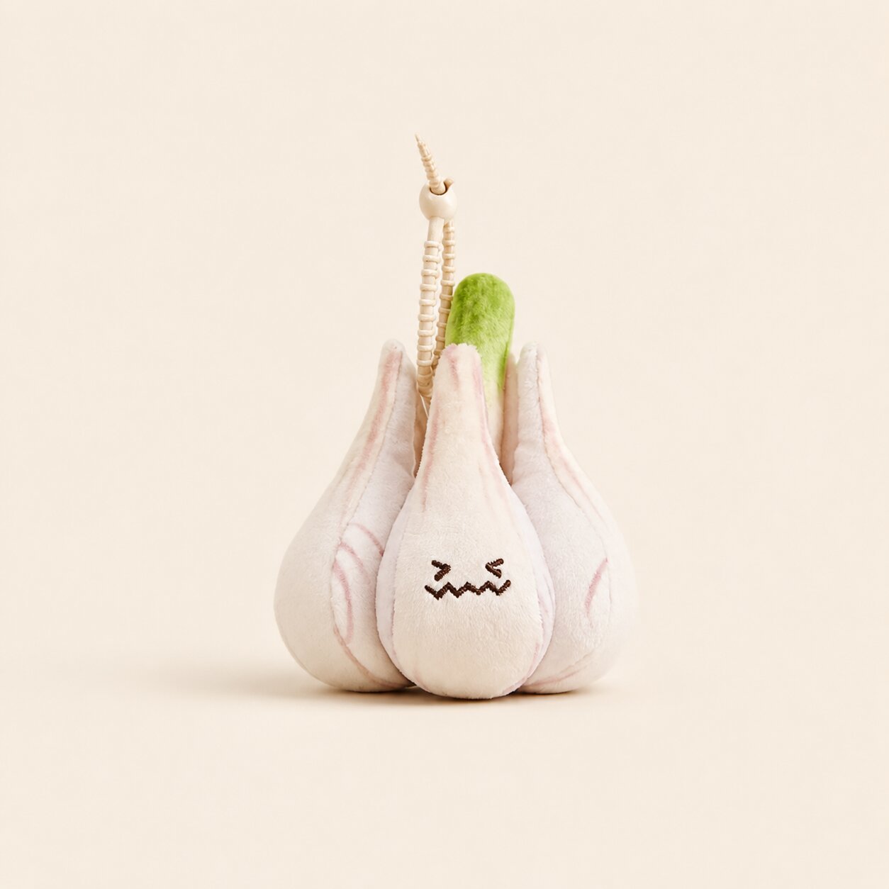 A plush garlic toy with a stitched frowning face, featuring pale pink and off-white fabric cloves, a green stem, and a small wooden bead keychain attached at the top, set against a solid cream background.