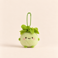 A cute, round green plush avocado keychain with embroidered facial features and a green ball chain, set against a solid pale peach background.