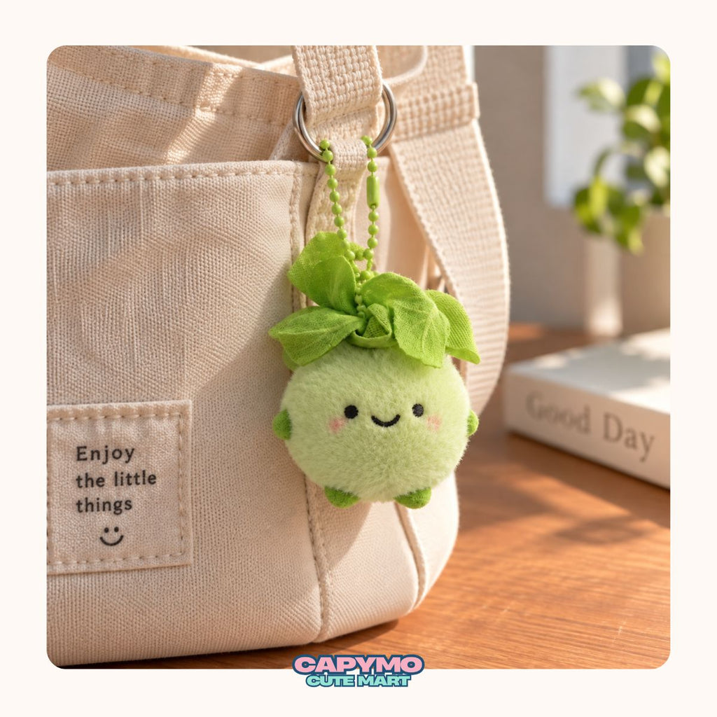 A cute green plush avocado keychain with a smiling face and leafy top, hanging from a beige canvas tote bag on a wooden table with a blurred book and plant in the background.