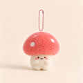 A cute mushroom-shaped plush keychain charm with a pinkish-red fuzzy cap, white polka dots, a smiling face, and a pink ball chain, shown on a warm beige background.