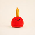 A cute red plush toy shaped like a small character with yellow cord-like hair, photographed against a plain warm off-white background.