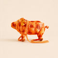 A small plush pig toy in orange-brown tones with an orange strap, photographed on a plain warm beige background.