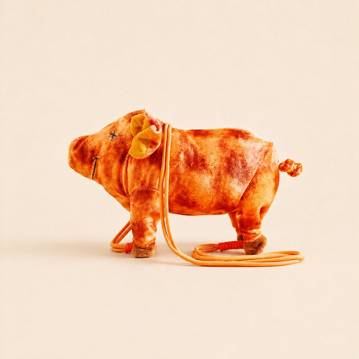 A small plush pig toy in orange-brown tones with an orange strap, photographed on a plain warm beige background.