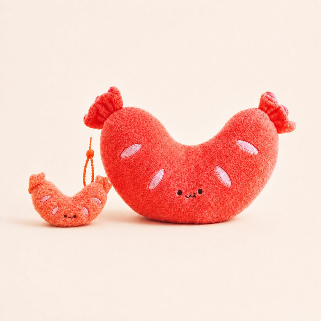 A plush sausage-shaped toy in bright red-orange with embroidered facial features and small pink oval details, shown next to a smaller matching plush accessory on a warm off-white background.