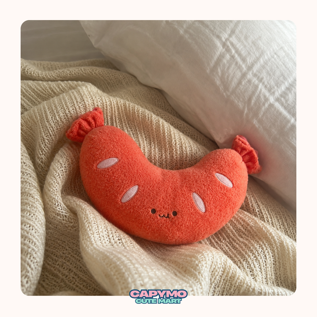 A plush sausage-shaped pillow in bright orange-red with embroidered white oval details and a small face, resting on a cream knit blanket next to a white pillow. A 'Cadymo Cute Mash' logo is visible at the bottom.