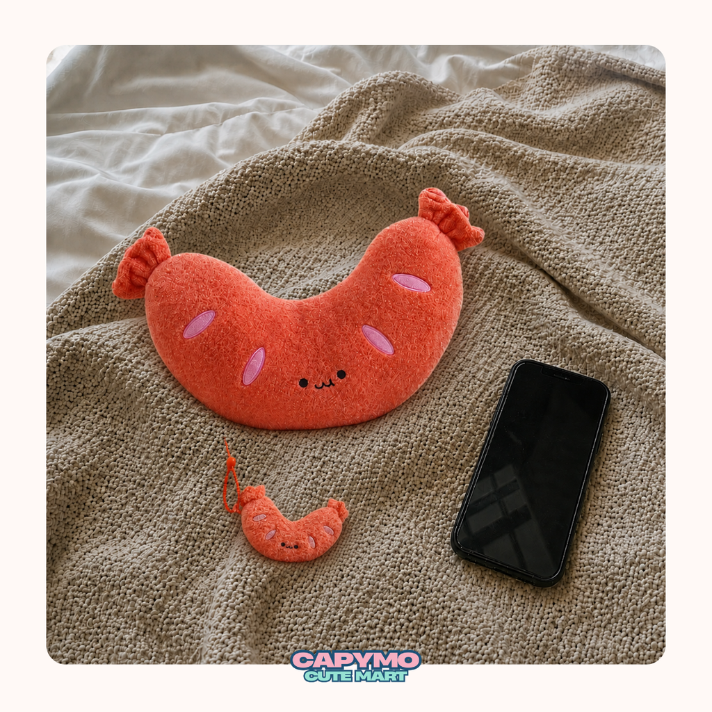 A plush sausage-shaped pillow in bright orange-red with pink cheeks and a small matching plush accessory, styled on a beige knit blanket next to a black smartphone. A 'Cadymo Cute Mash' logo is visible at the bottom.