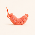 A small plush sausage-shaped keychain charm in a fuzzy coral-orange color, featuring embroidered facial features and a matching orange beaded loop, set against a solid off-white background.
