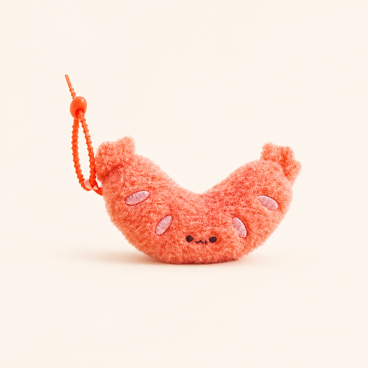 A small plush sausage-shaped keychain charm in a fuzzy coral-orange color, featuring embroidered facial features and a matching orange beaded loop, set against a solid off-white background.