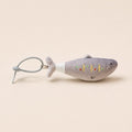 A small plush fish-shaped keychain or bag charm in light gray, with embroidered facial features and a white wrapped neck, attached to a gray metal ring on a plain beige background.