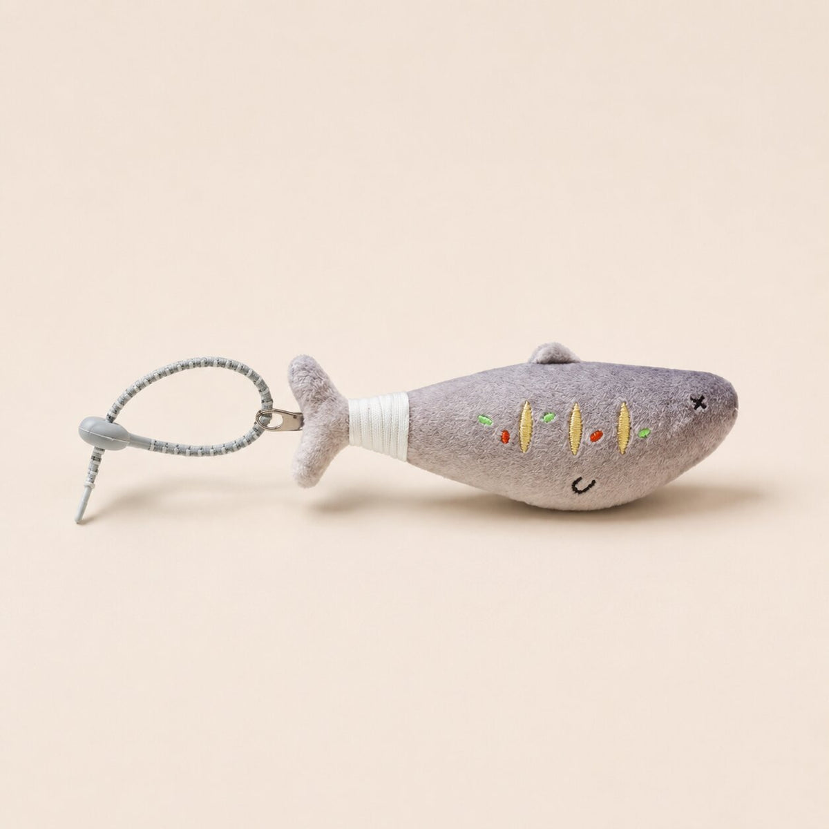 A small plush fish-shaped keychain or bag charm in light gray, with embroidered facial features and a white wrapped neck, attached to a gray metal ring on a plain beige background.