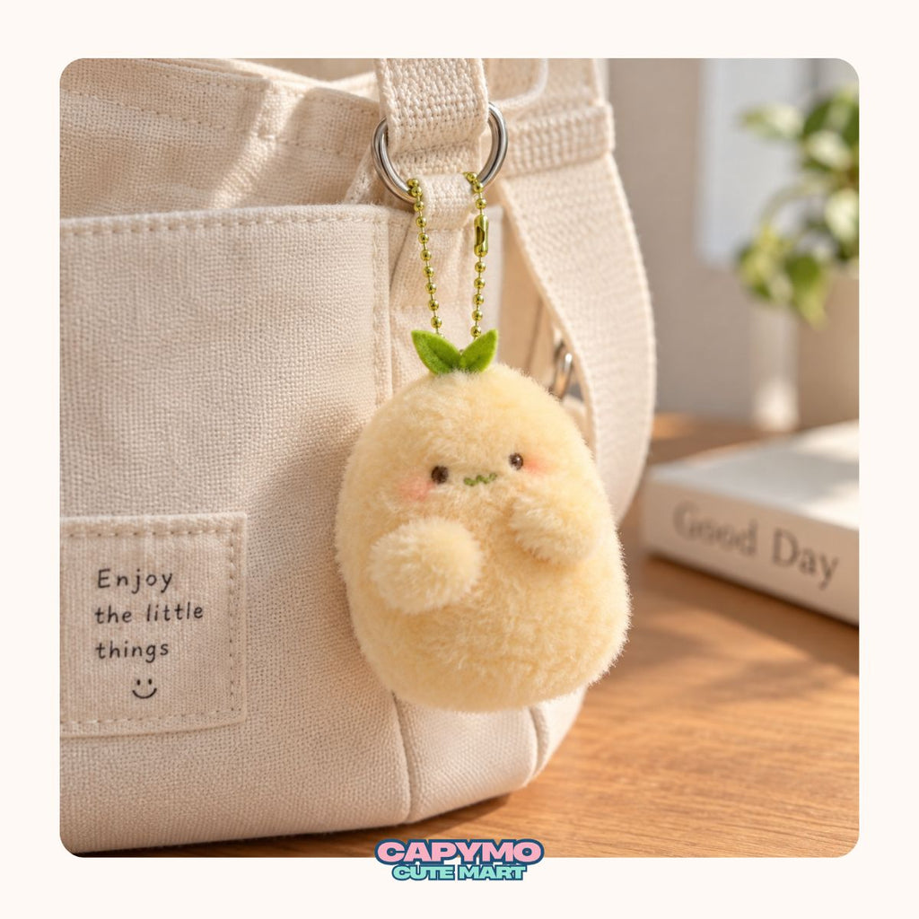 A cute, round, pale yellow plush character keychain with a small green leaf on its head, hanging from a silver ring on a beige canvas tote bag. The background shows a warm, softly blurred indoor setting with a wooden table and a book.