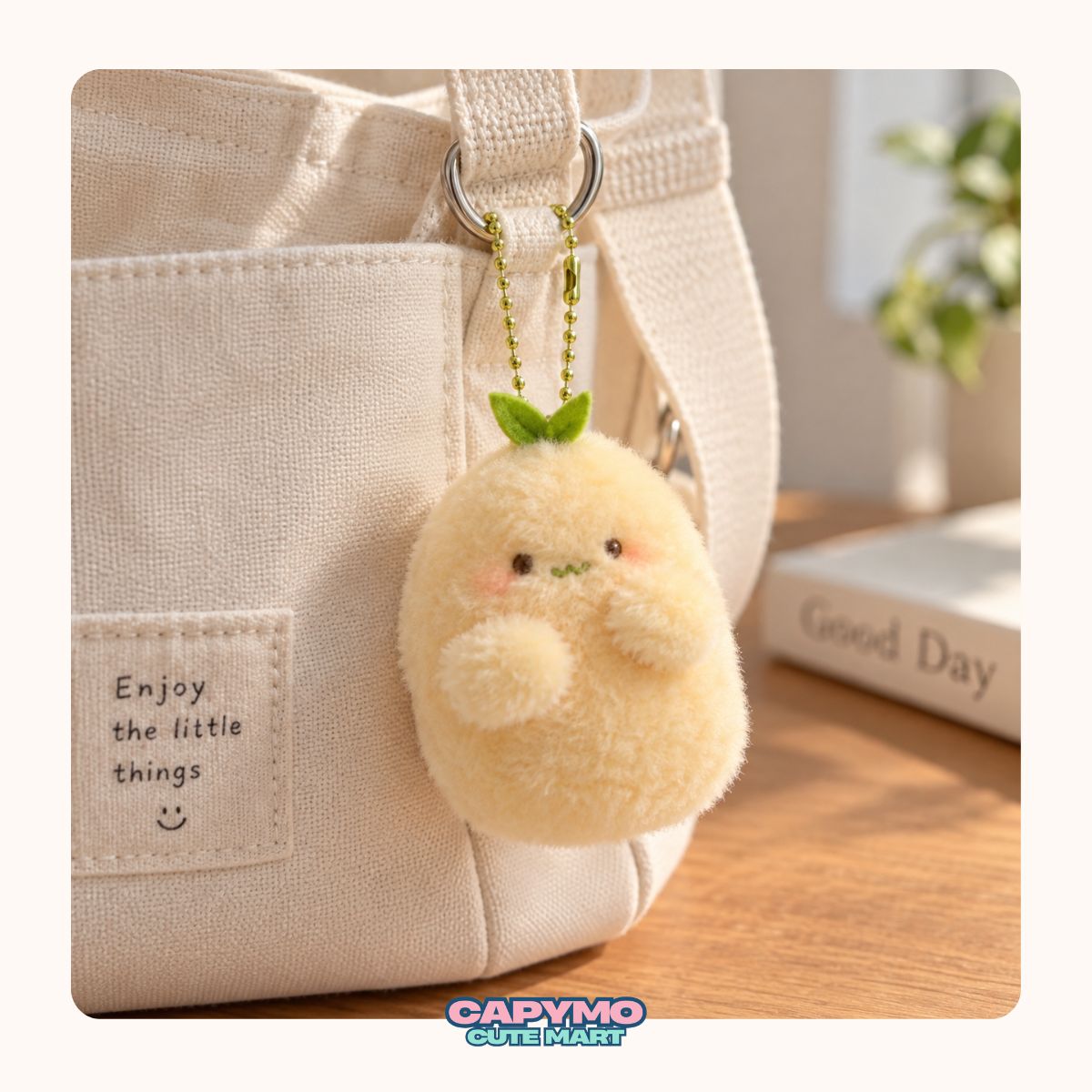 A cute, round, pale yellow plush character keychain with a small green leaf on its head, hanging from a silver ring on a beige canvas tote bag. The background shows a warm, softly blurred indoor setting with a wooden table and a book.