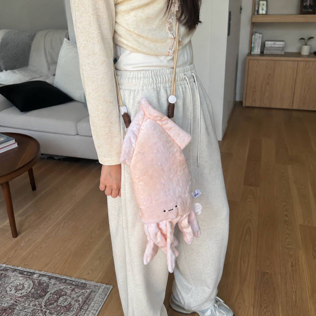 A person wearing a cream ribbed long-sleeve top and cream sweatpants carries a plush pink octopus-shaped crossbody bag with a wooden bead strap in a modern living room setting.