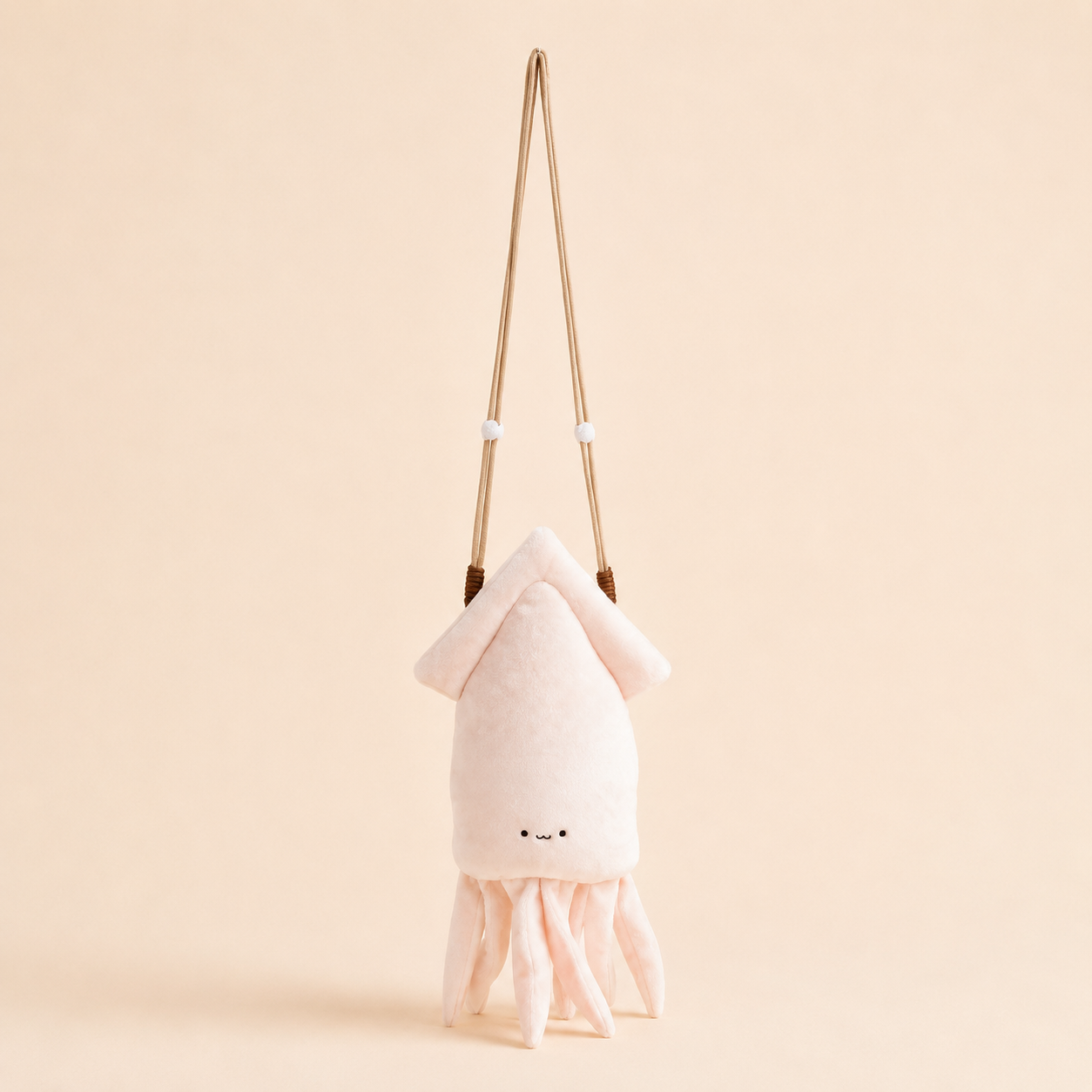 A plush octopus-shaped shoulder bag in pale pink and cream, featuring a long tan strap and a small embroidered face, set against a solid light beige background.