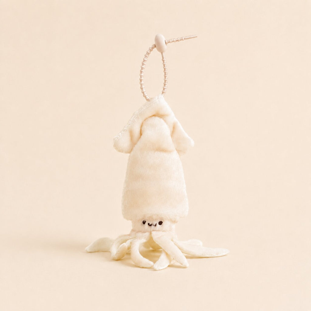 A small plush octopus or squid character keychain in warm off-white/cream, hanging from a thin beaded loop on a plain beige background.