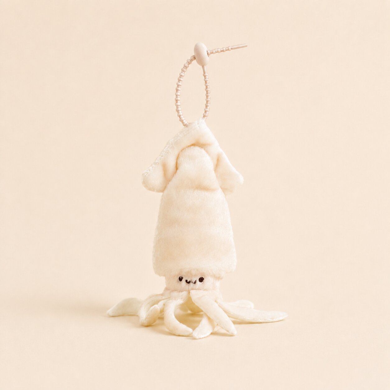 A small plush octopus or squid character keychain in warm off-white/cream, hanging from a thin beaded loop on a plain beige background.