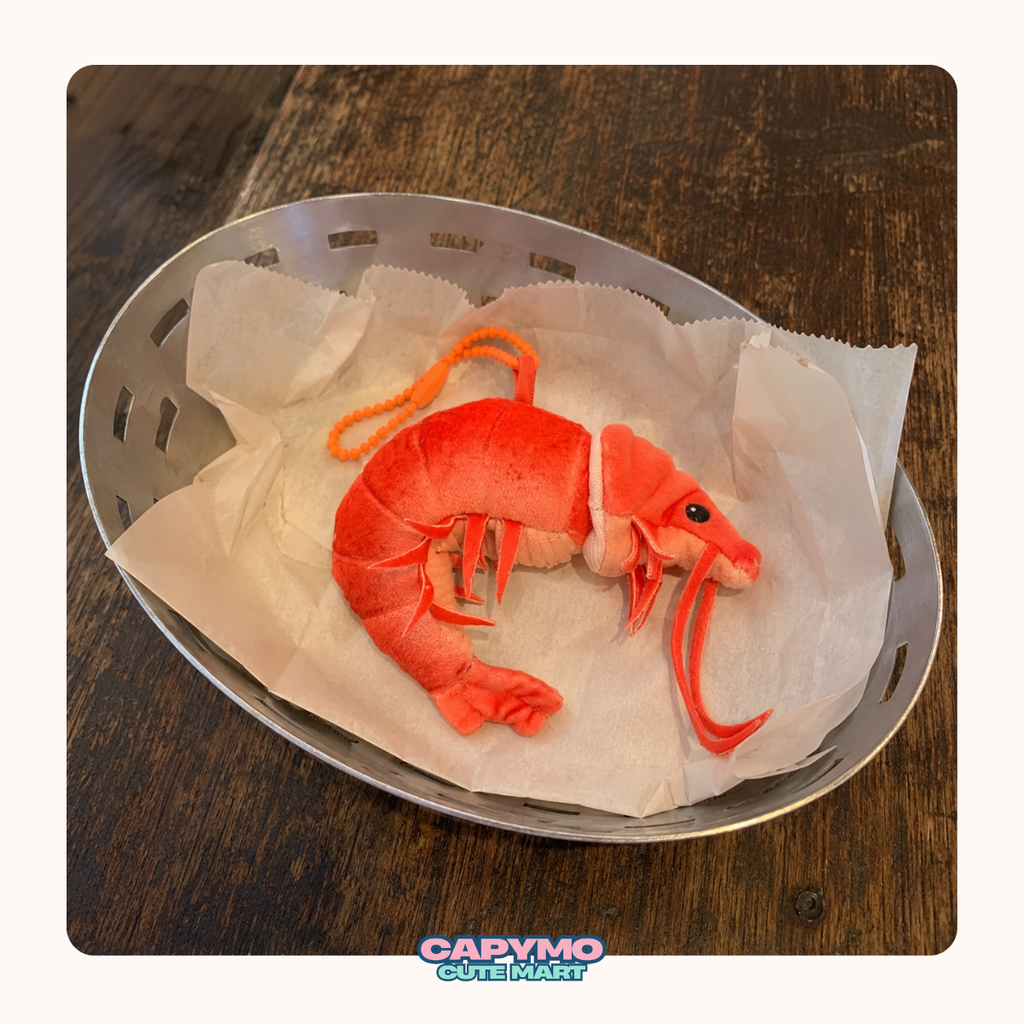 A plush toy shrimp or prawn in bright orange-red and pink tones, placed on white tissue paper inside a metal oval basket on a dark wooden table. A 'Capymo Cute Market' logo is visible at the bottom.