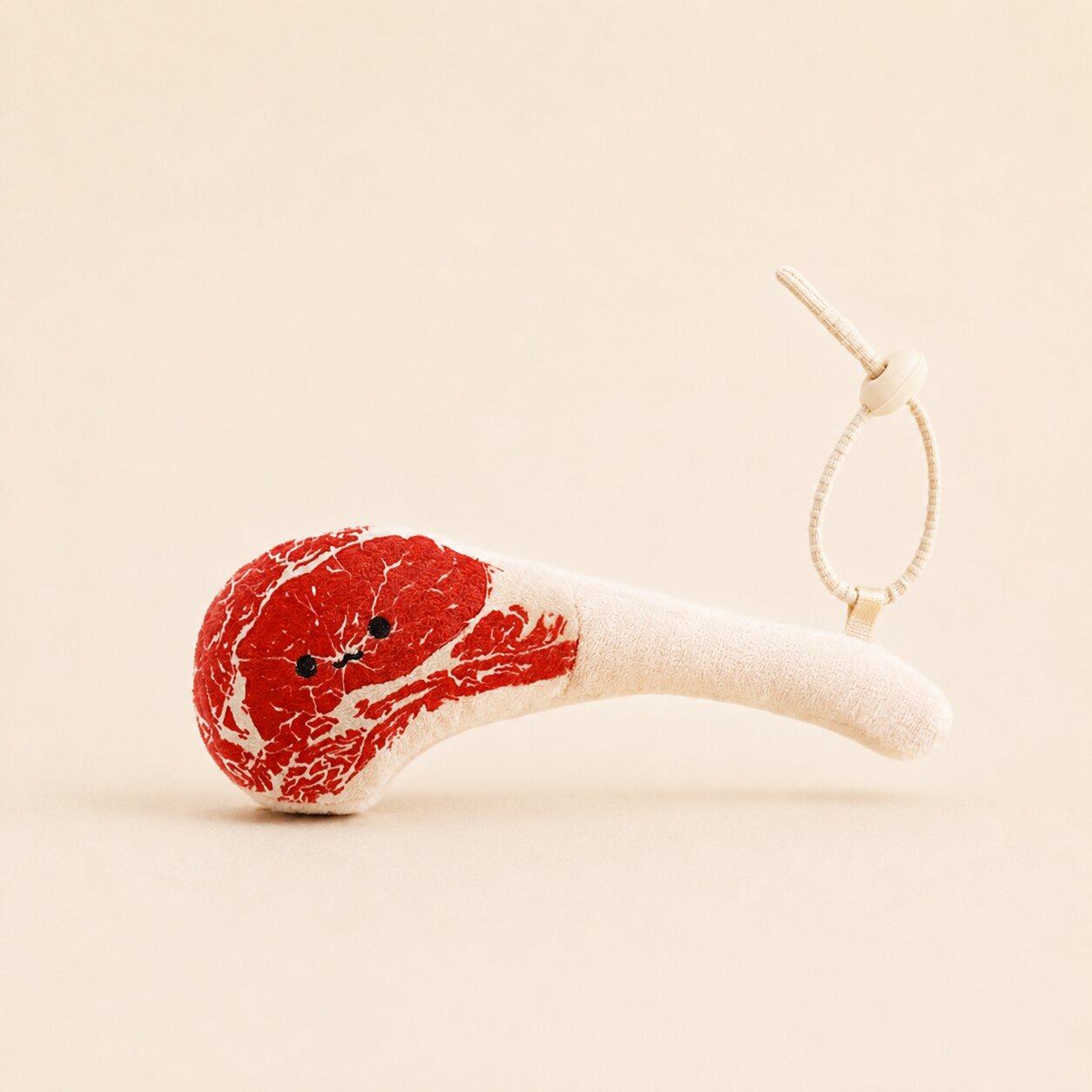 A plush toy shaped like a raw meat cut with red marbled texture and a cream-colored stem, featuring small black embroidered eyes and a cream hanging loop on a warm beige background.