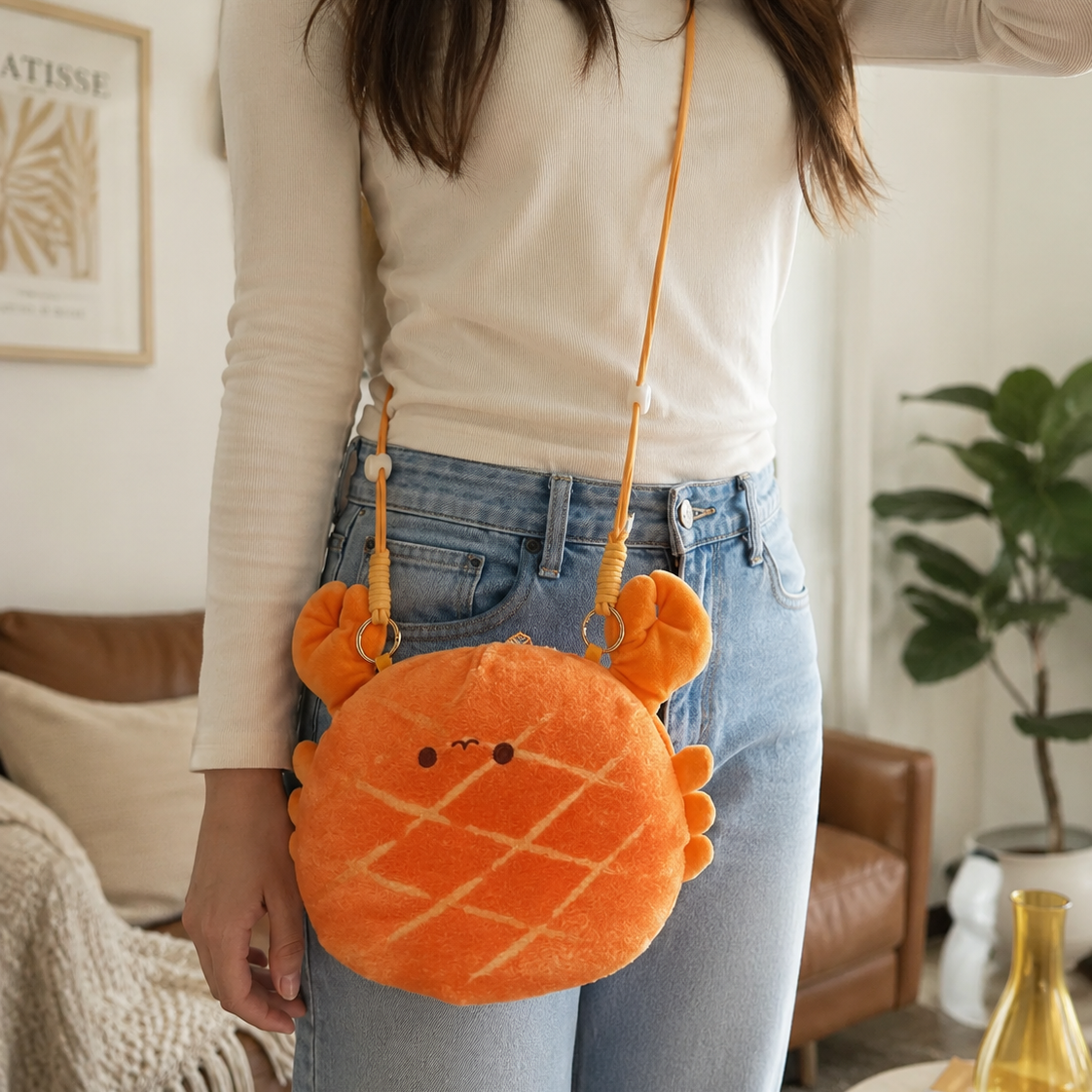 A person wearing a cream long-sleeve top and light blue jeans carries a round orange plush crossbody bag shaped like a crab, with a thin orange strap and small embroidered facial features.