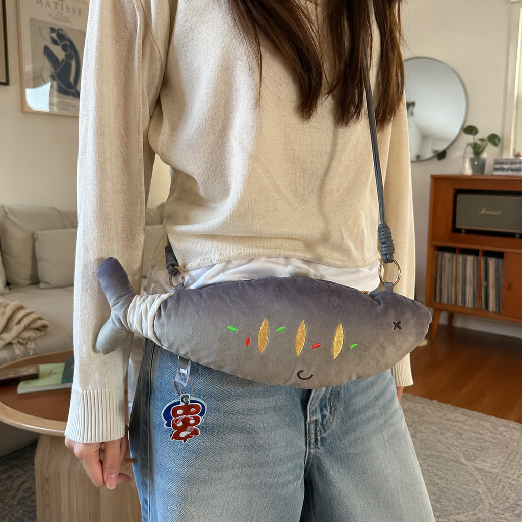 A person wearing a cream knit sweater and light blue jeans carries a gray plush fish-shaped crossbody bag with embroidered details in a cozy living room setting.