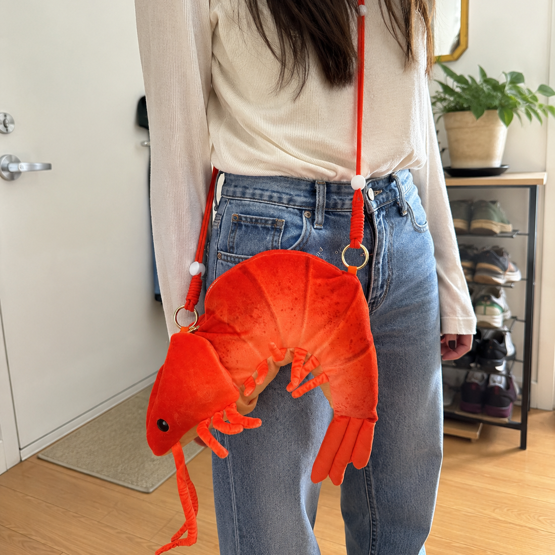A person wearing a cream long-sleeve top and light blue jeans carries a bright orange lobster-shaped crossbody bag with a red strap in an indoor room setting.