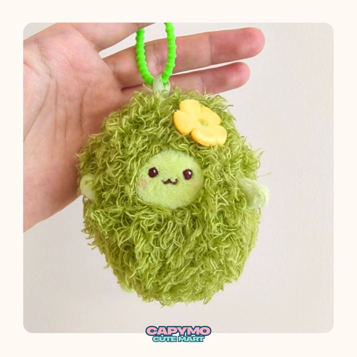 Green plush keychain with a face held in a hand against a white background, featuring the brand 'Capymo Cute Mart'.