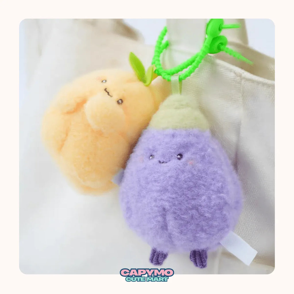 Two plush keychains, one orange and one purple, with a green clip on a white background. Brand 'Capymo' is visible.